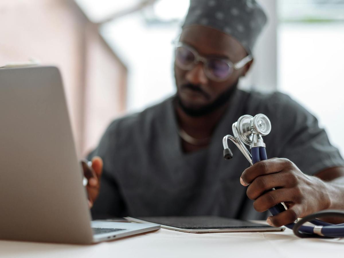 a man in gray scrub suit holding stethoscope while typing on his laptop